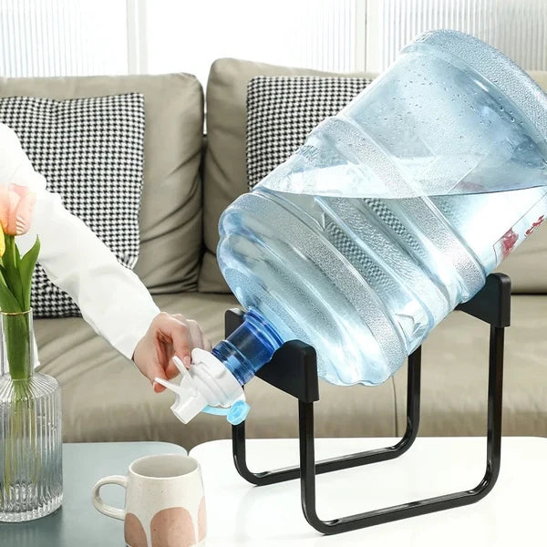 Person pouring water from a large plastic bottle into a cup on a table with a couch in the background.