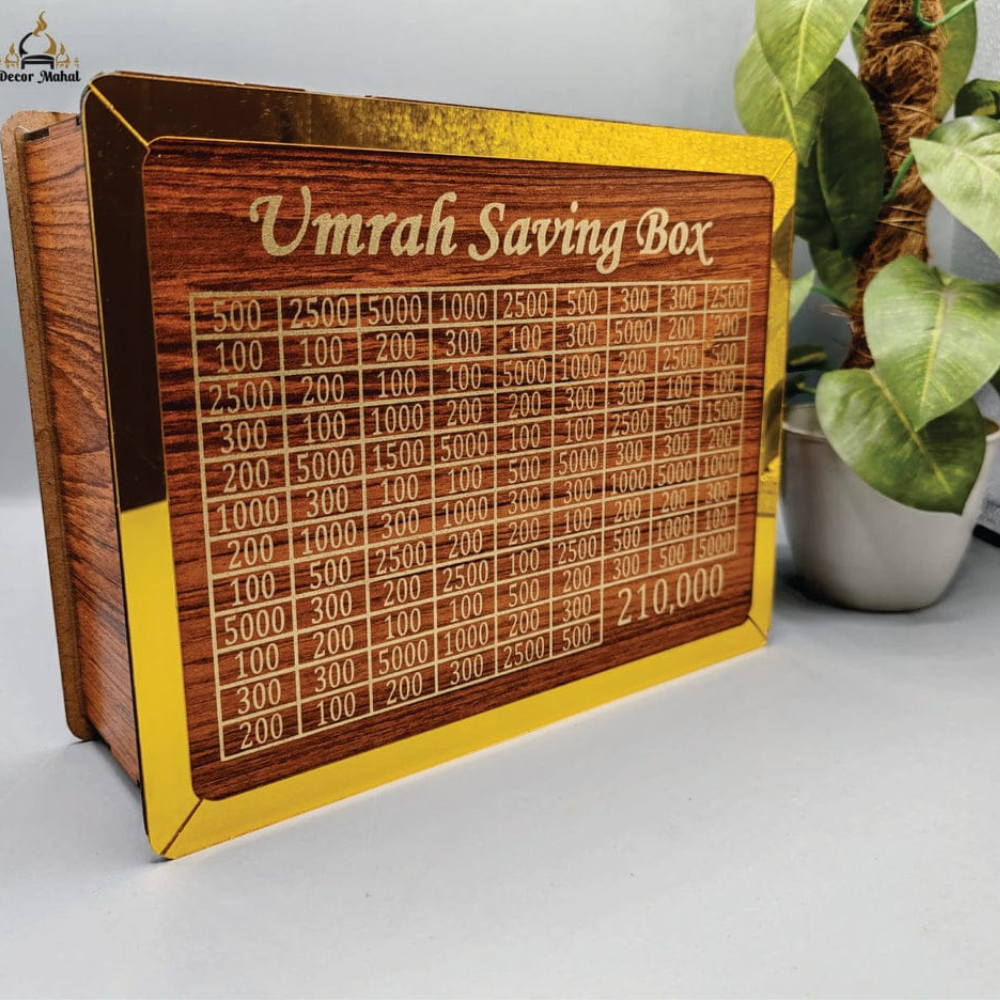 A wooden Umrah saving box with a gold-colored frame and engraved text showing a progress table, displayed on a light gray surface next to a potted plant.