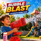 A young boy smiling and holding a red automatic bubble gun toy that is spraying bubbles into the air. The scene is set in a backyard with a playground visible in the background under a blue sky.