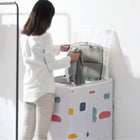 A woman in a white shirt and beige pants stands beside a compact top-loading washing machine covered with a white fabric dust cover decorated with colorful abstract shapes. She is holding a gray towel near the open lid.