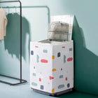 A top-loading washing machine covered with a white fabric dust cover decorated with colorful abstract geometric shapes, set against a teal wall with a clothing rack in the background.